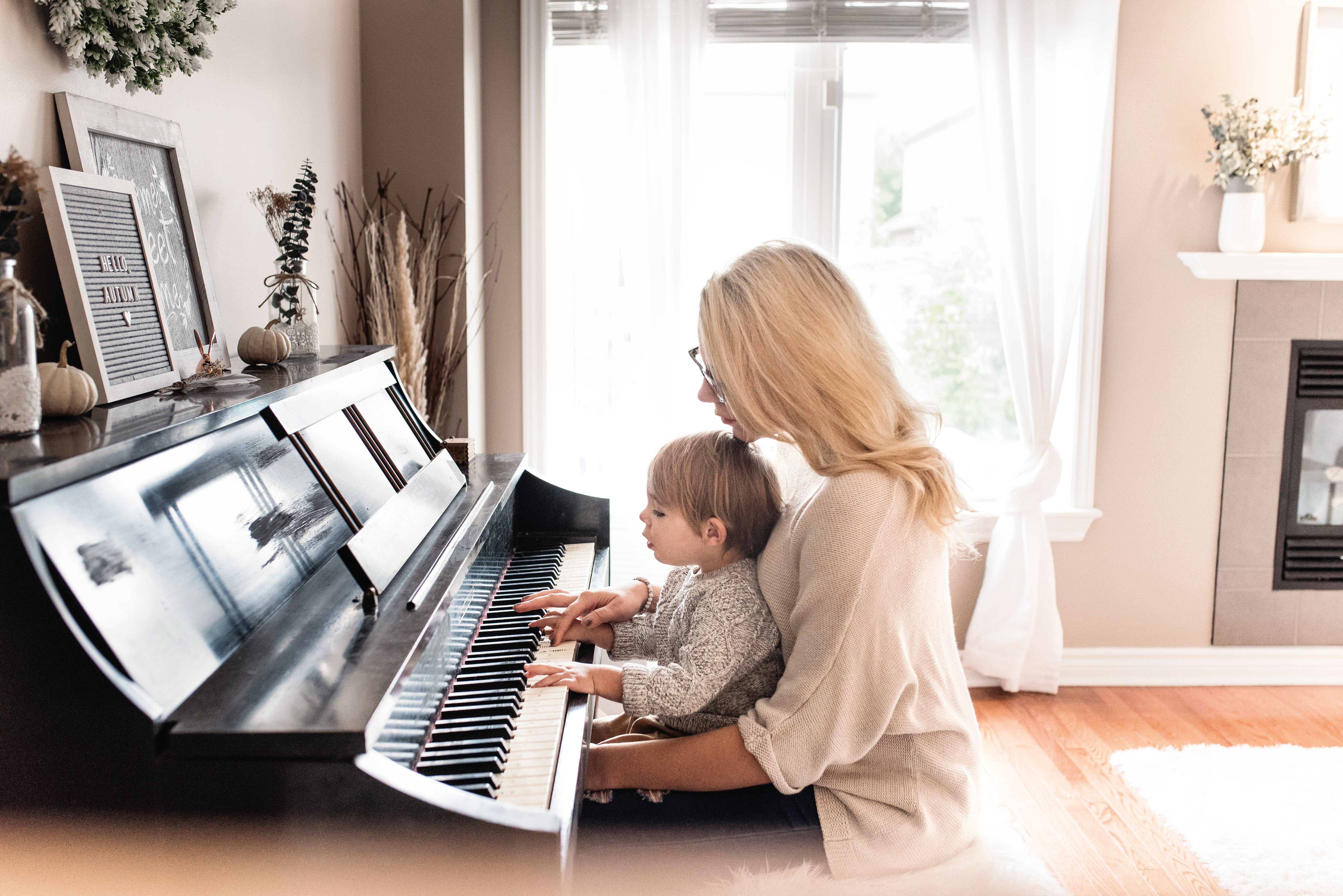 young boy sits on a woman's lap at an upright piano and plays from sheet music